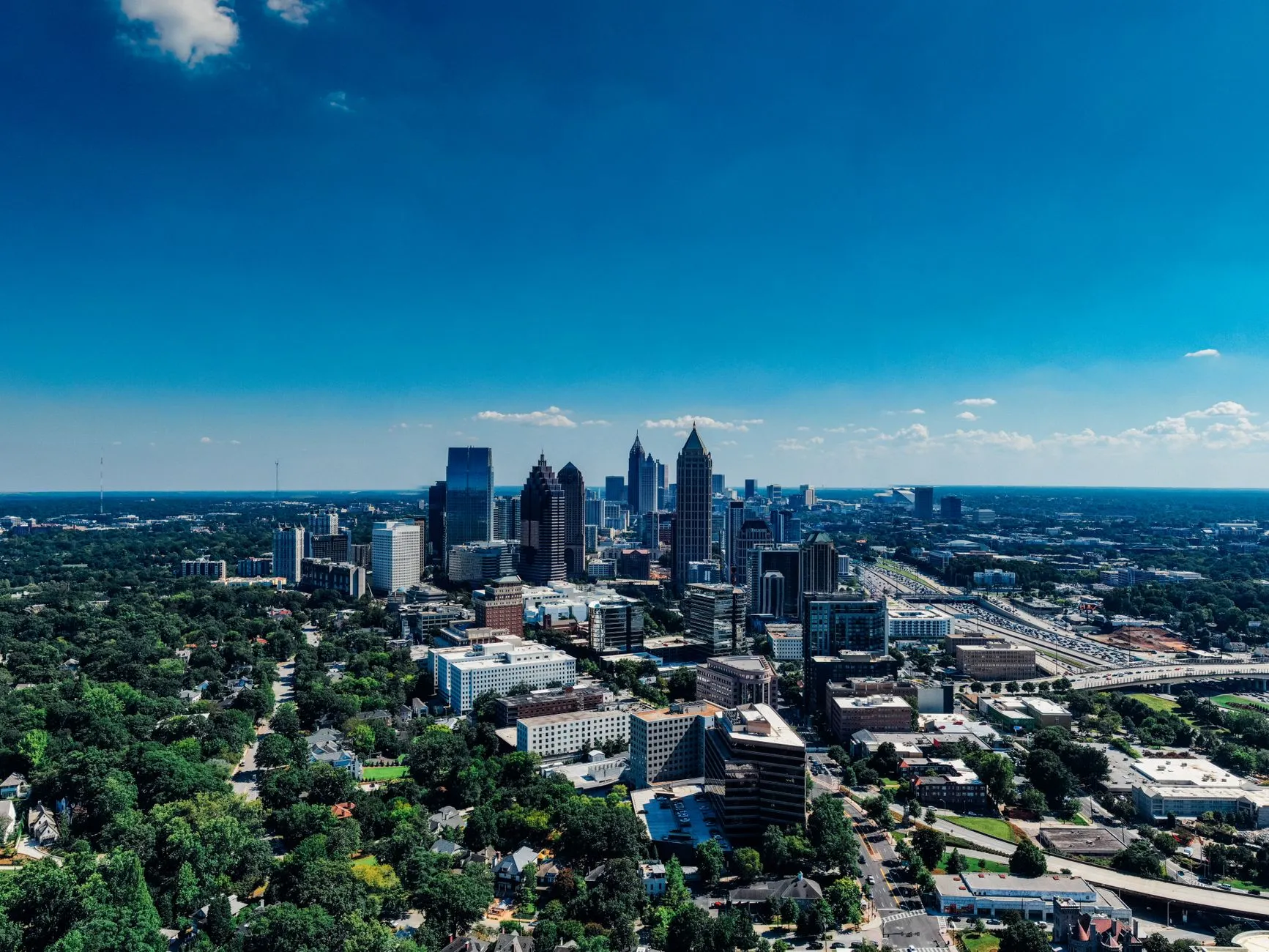 Midtown Atlanta from above under a deep blue sky, with the pyramid-crowned One Atlantic Center and the gold spire of Bank of America Plaza rising out of the city's tree canopy.