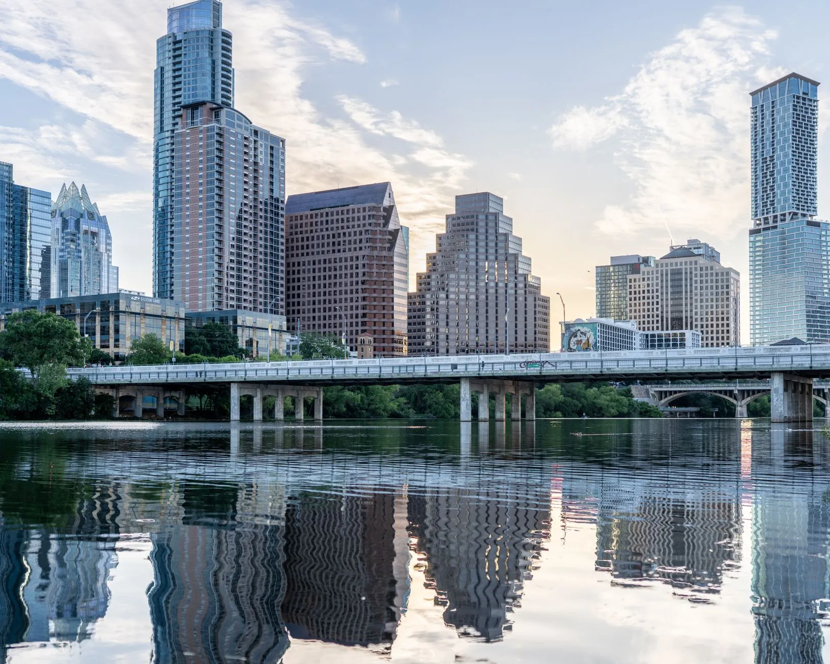 The Austin skyline reflected in Lady Bird Lake at first light, with the Frost Bank Tower's faceted crown on the left and the Congress Avenue Bridge running across the water.