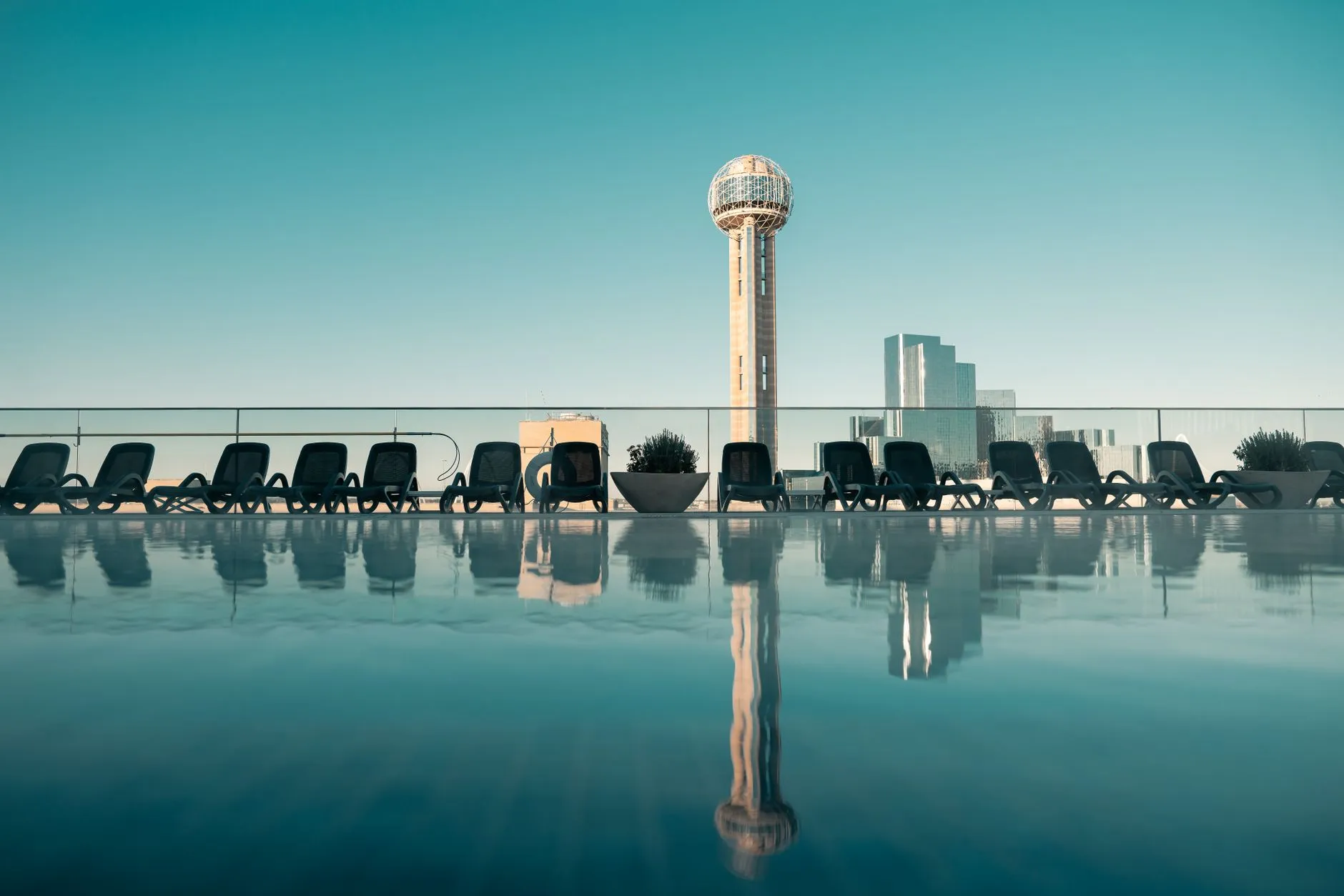 Reunion Tower and the downtown Dallas skyline standing against a clear sky and mirrored in the still water of a rooftop pool.