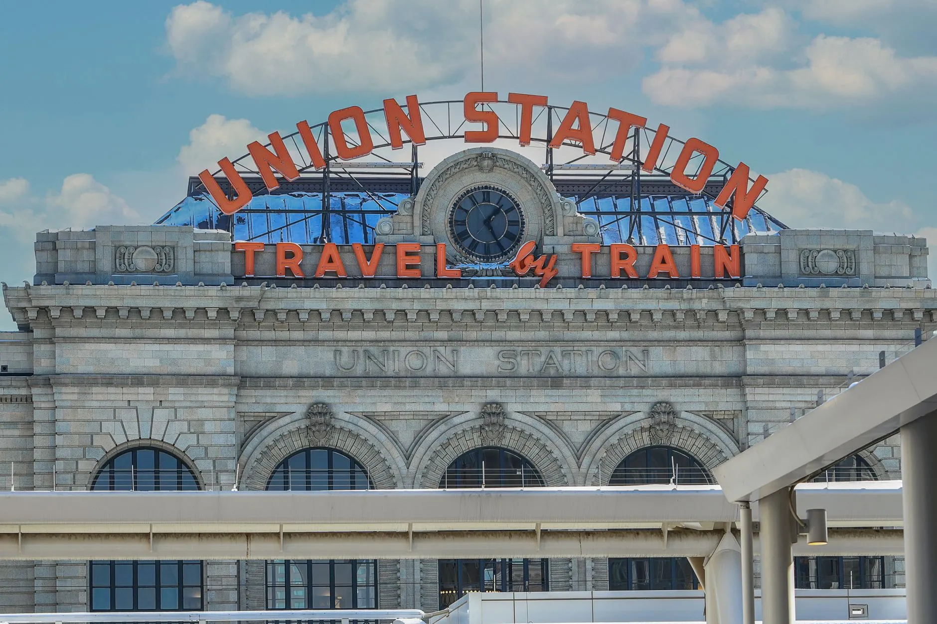 The front of Denver Union Station under a blue sky, its carved granite facade topped by the red neon arch reading Union Station and Travel by Train.