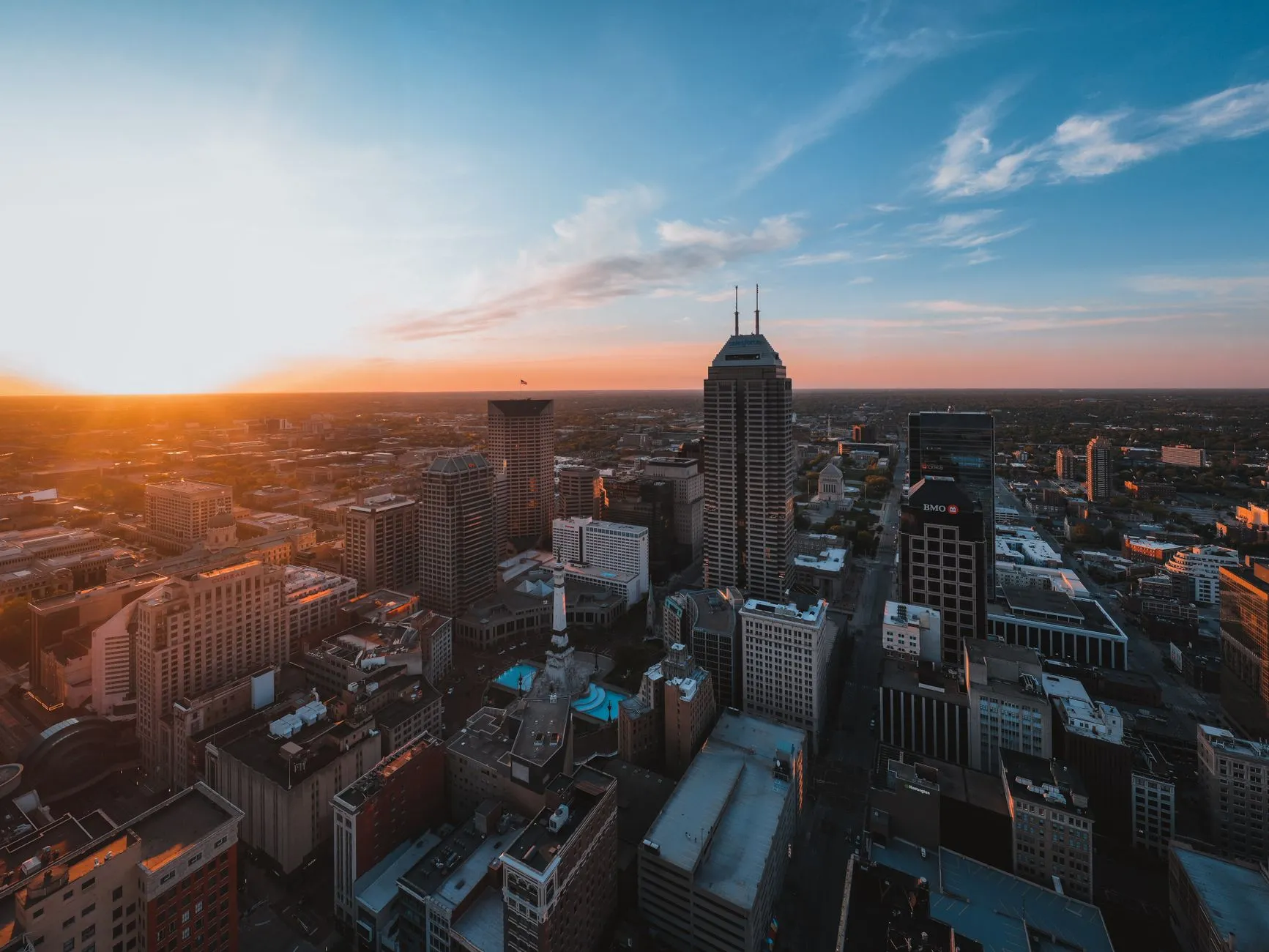 Downtown Indianapolis from the air in late afternoon light, with the Soldiers and Sailors Monument and its fountains at the center of Monument Circle and Salesforce Tower rising beside it.