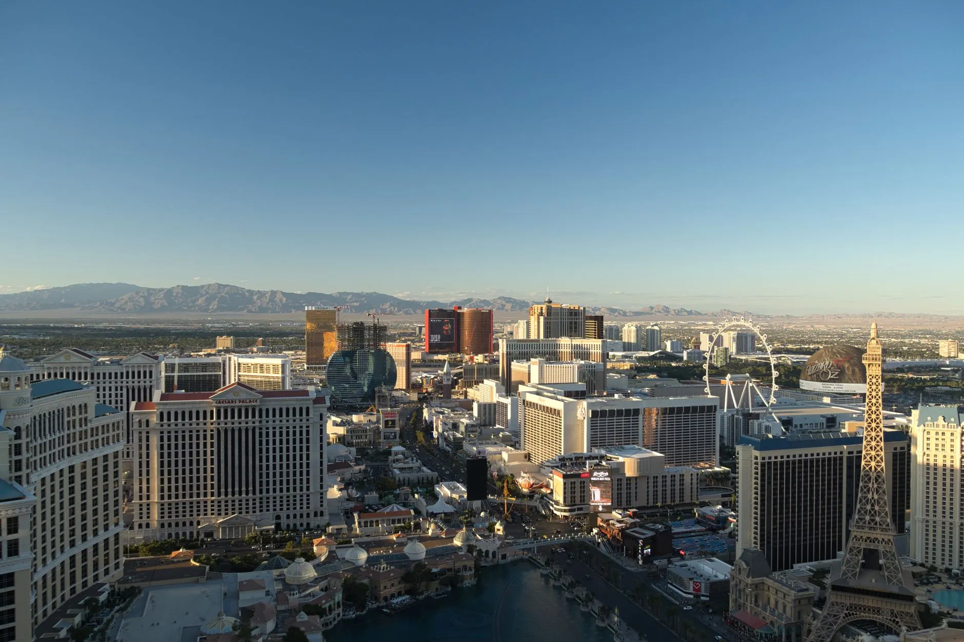 The Las Vegas Strip from above in early light, with Caesars Palace, the Bellagio fountain lake, the half-scale Eiffel Tower at Paris Las Vegas and the High Roller wheel set against the desert mountains.
