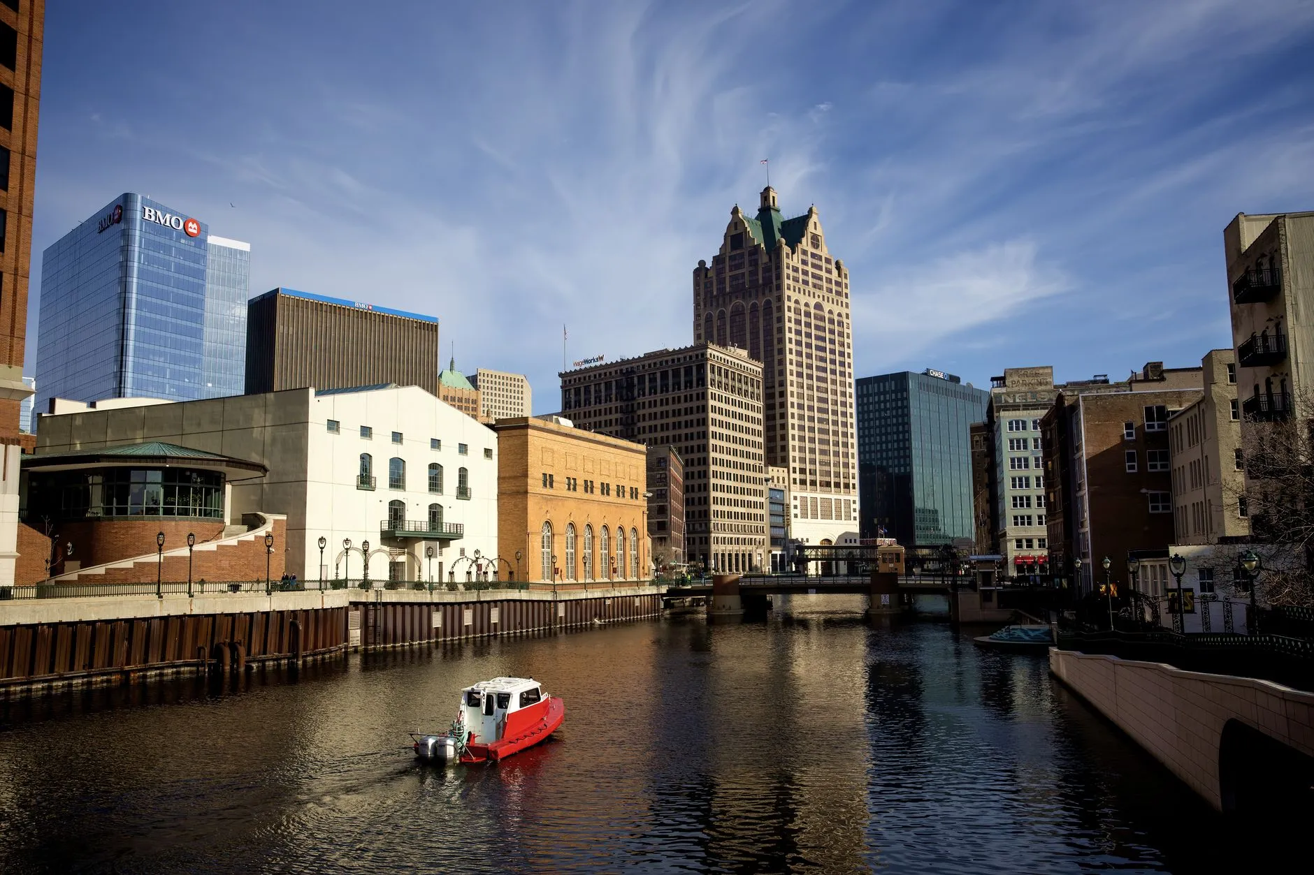 A small boat on the Milwaukee River downtown, with the gabled crown of 100 East Wisconsin rising over the Riverwalk on a clear day