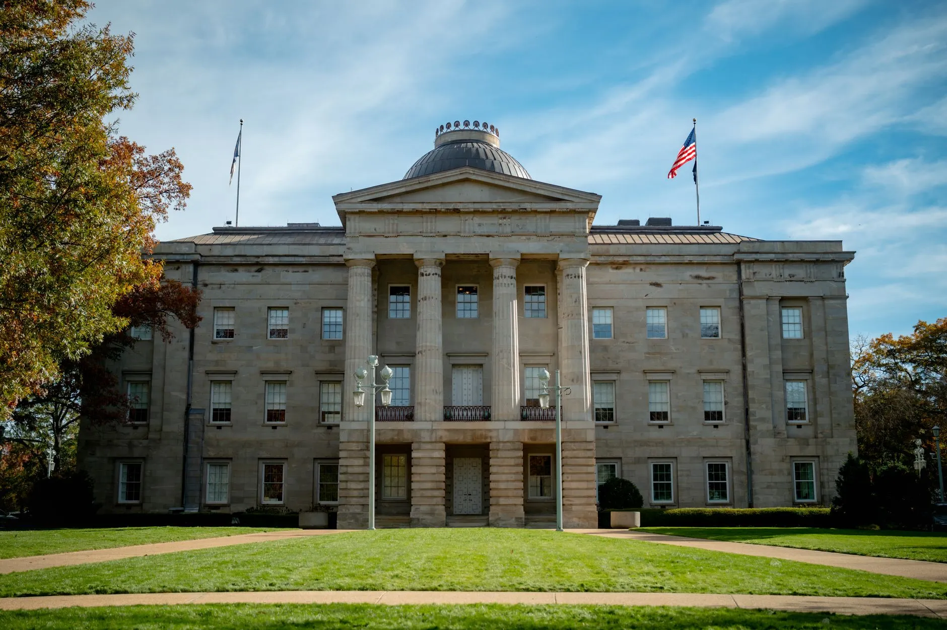The North Carolina State Capitol in Raleigh on a bright autumn day, its Doric portico and low domed rotunda framed by turning trees and flags.